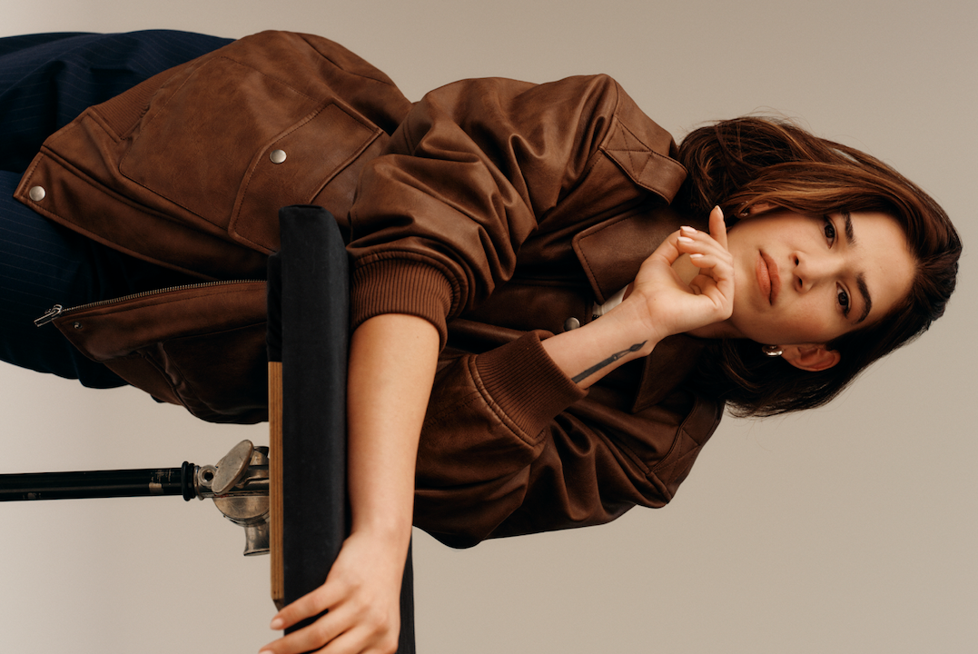 H&M employee in a brown leather jacket leaning on a stool, facing the camera.