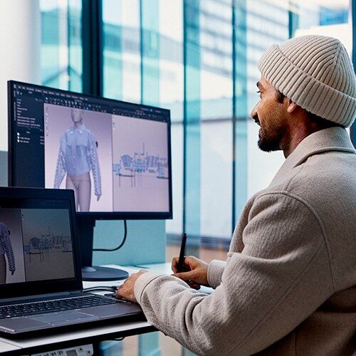 一名男性坐在电脑前，你在电脑屏幕上看到一件夹克的3D设计。